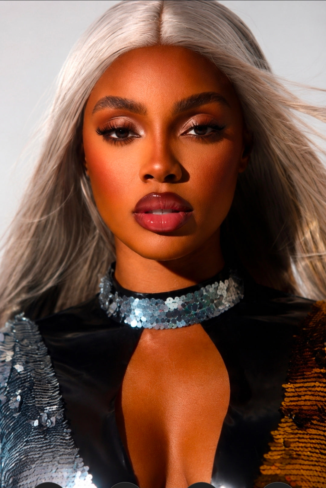 A close-up portrait of a model with long platinum blonde hair, wearing a black sequined top with a silver choker collar and textured metallic side panels.