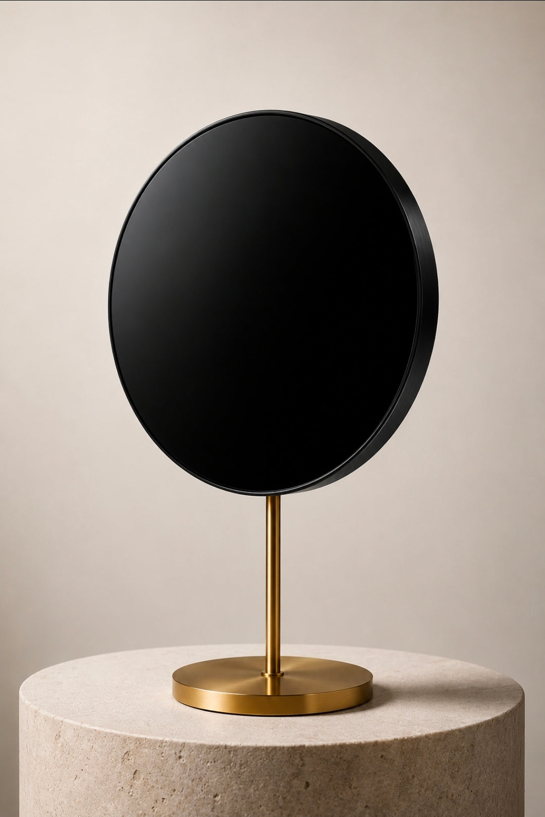 A round black mirror with a slim gold stand and circular gold base, displayed on a round stone pedestal against a warm beige background.